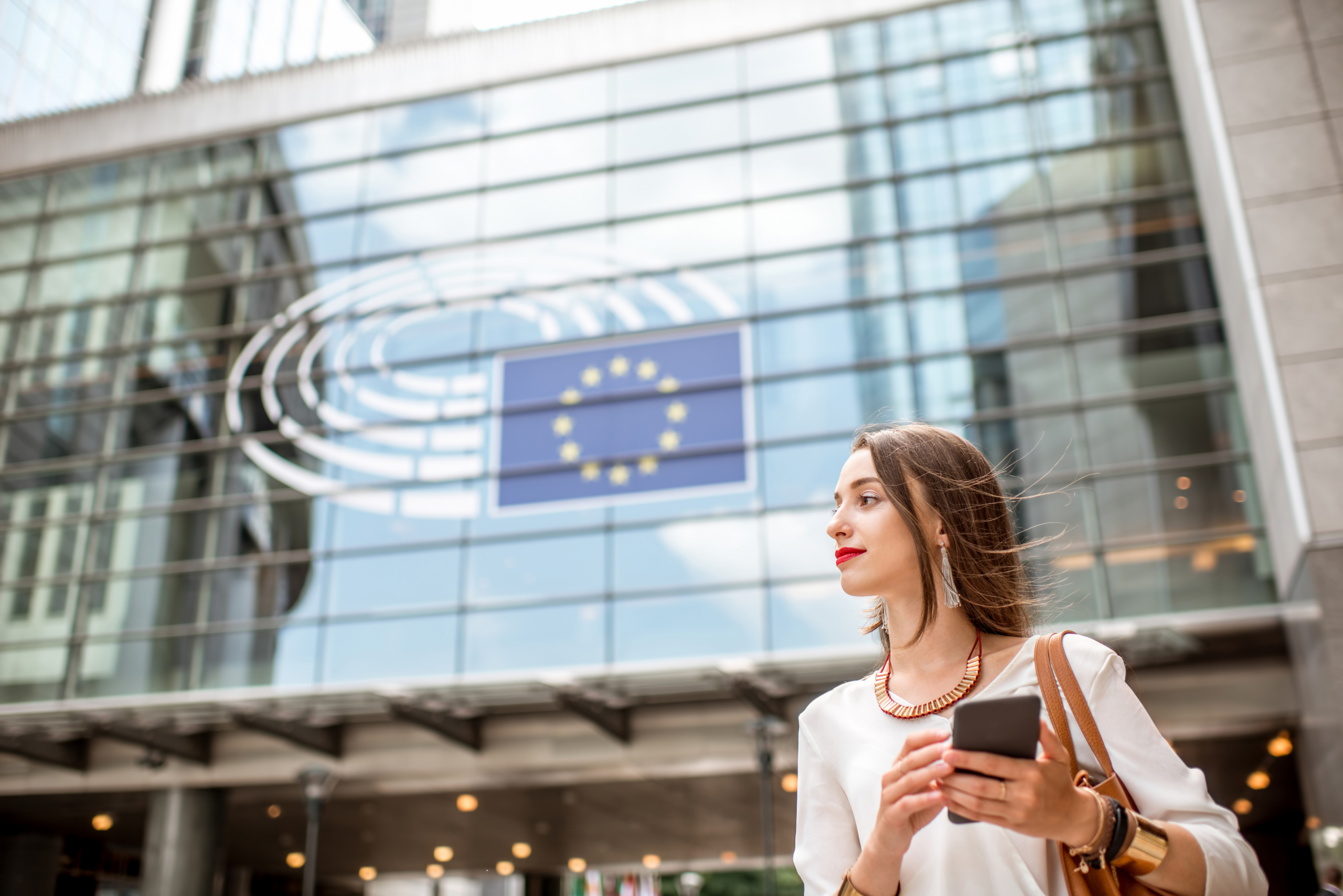 Frau in elegantem Business-Outfint steht vor Glasfront eines EU-Gebäudes.