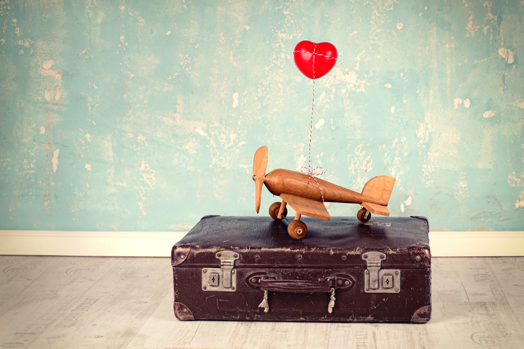 Holzflugzeug auf altem Lederreisekoffer. Am Flugzeug ist ei Herzballon befestigt. Sprache für den Urlaub